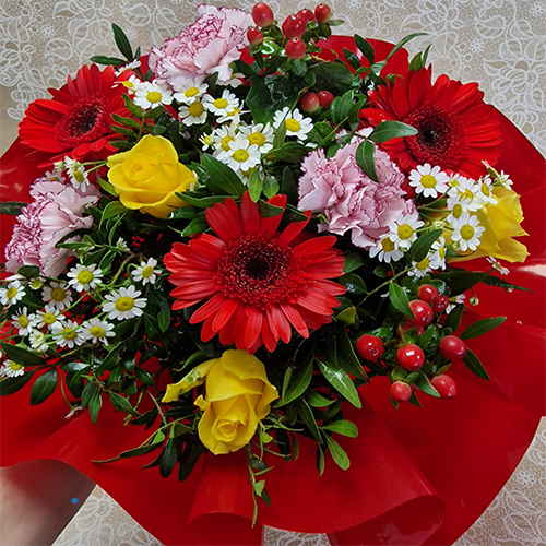 Bouquet of red gerbera daisies, yellow roses, pink carnations, and white daisies with green leaves.